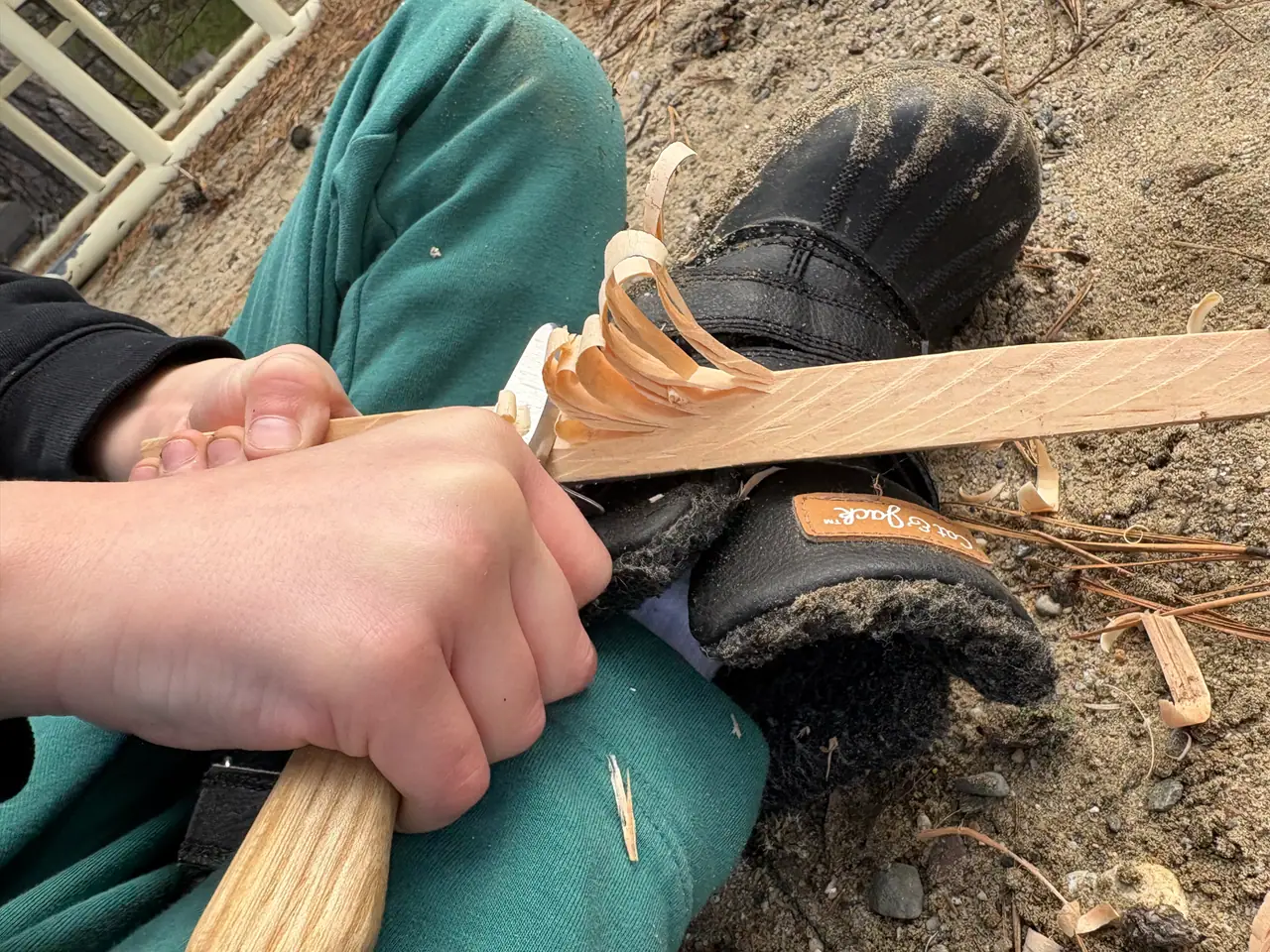 Small hands carve a stick with a fixed-blade knife, shavings curling off the wood.
